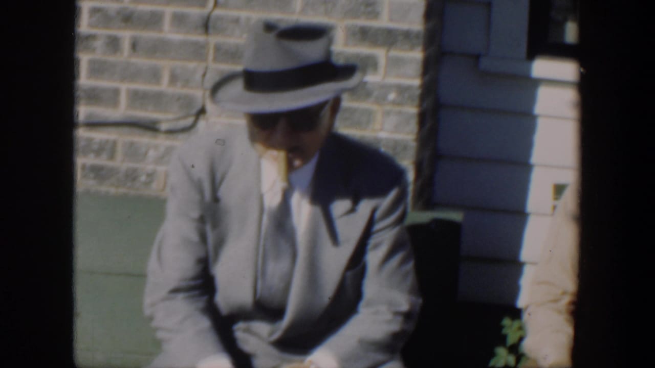 Older man in a gray suit and fedora sits outdoors beside a stone wall.