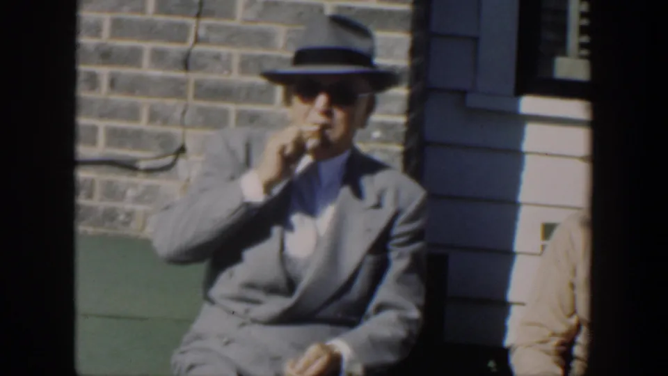1951 Cuba home-movie frame showing the seated man in a suit and fedora raising a cigar near a stone wall.
