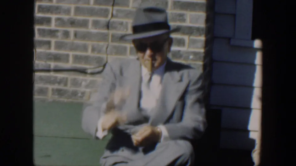 1951 Cuba frame of the seated man in a gray suit and fedora gesturing with a cigar beside a stone wall and green ledge.