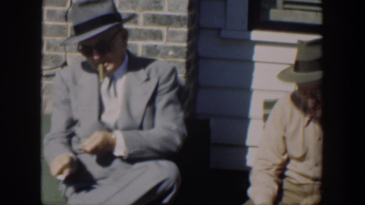 Man in a gray suit and fedora sits on a green bench beside another seated man.