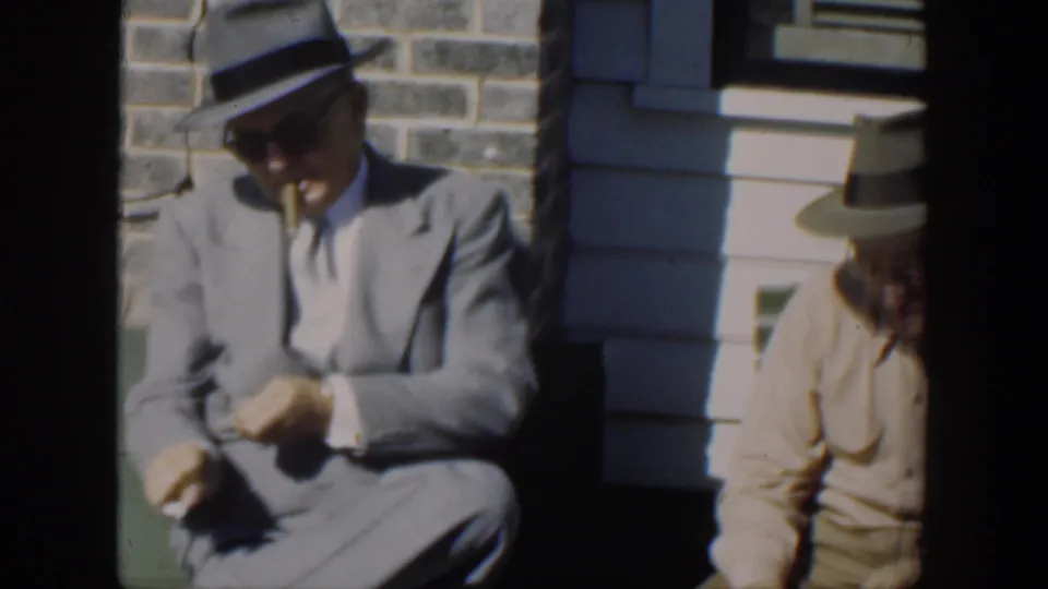 In 1951 Cuba home-movie footage, a suited man in a fedora sits in sunlight on a green bench near another man in a hat.