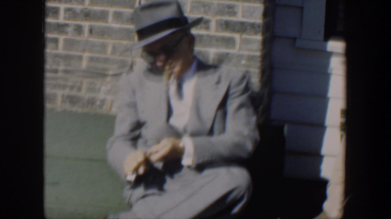 Suited man in fedora sits alone in frame, looking down at his hands.