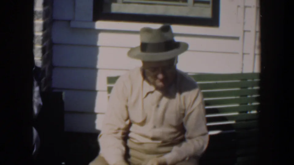 A second seated man in a light shirt and brimmed hat appears on the green bench outside the brick and sided building.