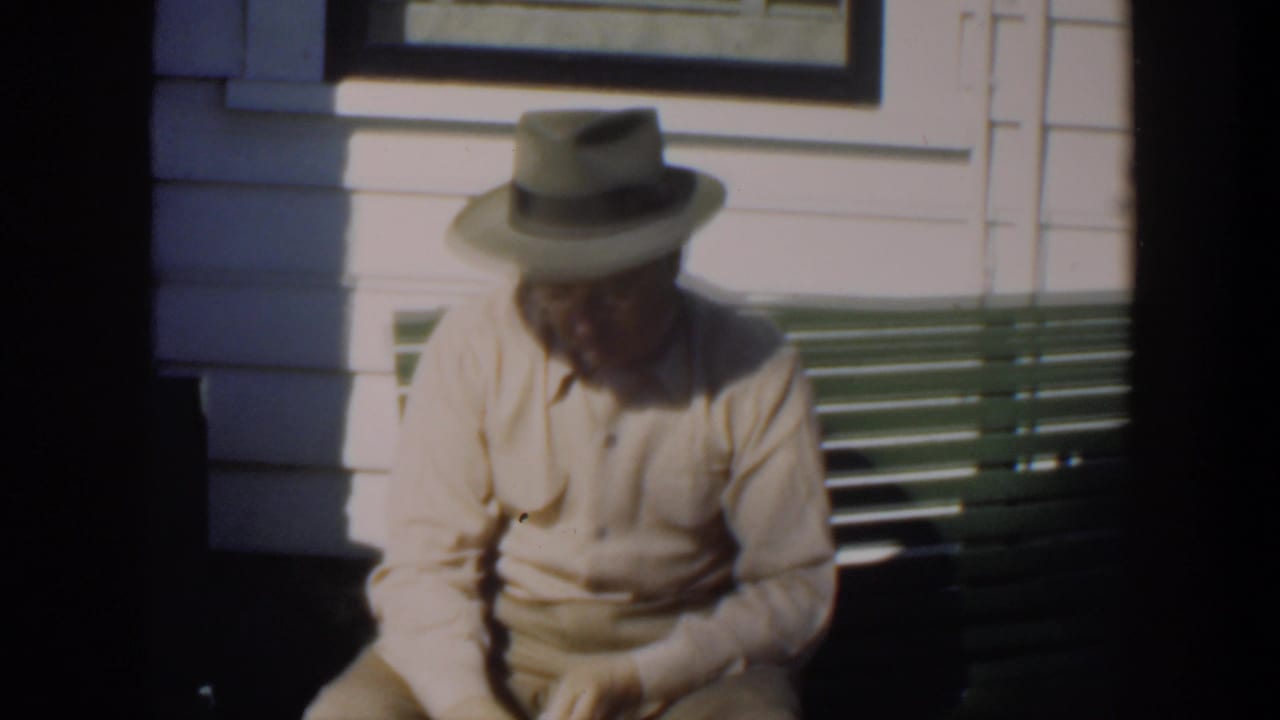 Elderly man in a fedora sitting on a green bench outside a white clapboard wall.