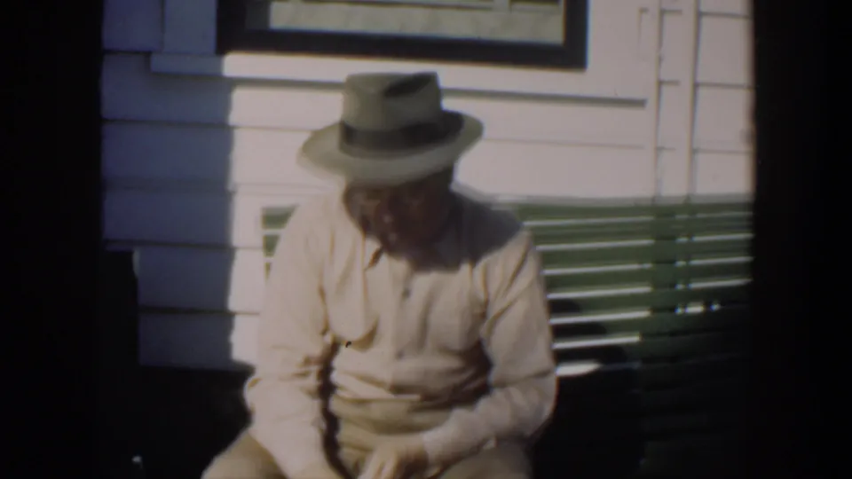 In Cuba home-movie footage from 1951, an elderly man in a light shirt and fedora sits on a green porch bench in strong sun.