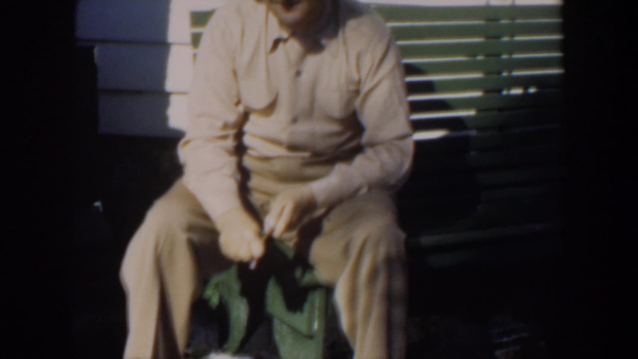 Seated older man cropped at the shoulders, holding a small object near a green bench.