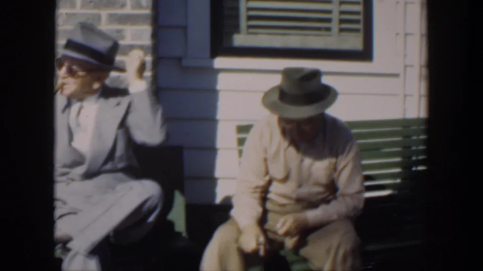 Two older men sit on the porch bench in 1951 Cuba footage, one in a suit and hat and the other looking down at his hands.