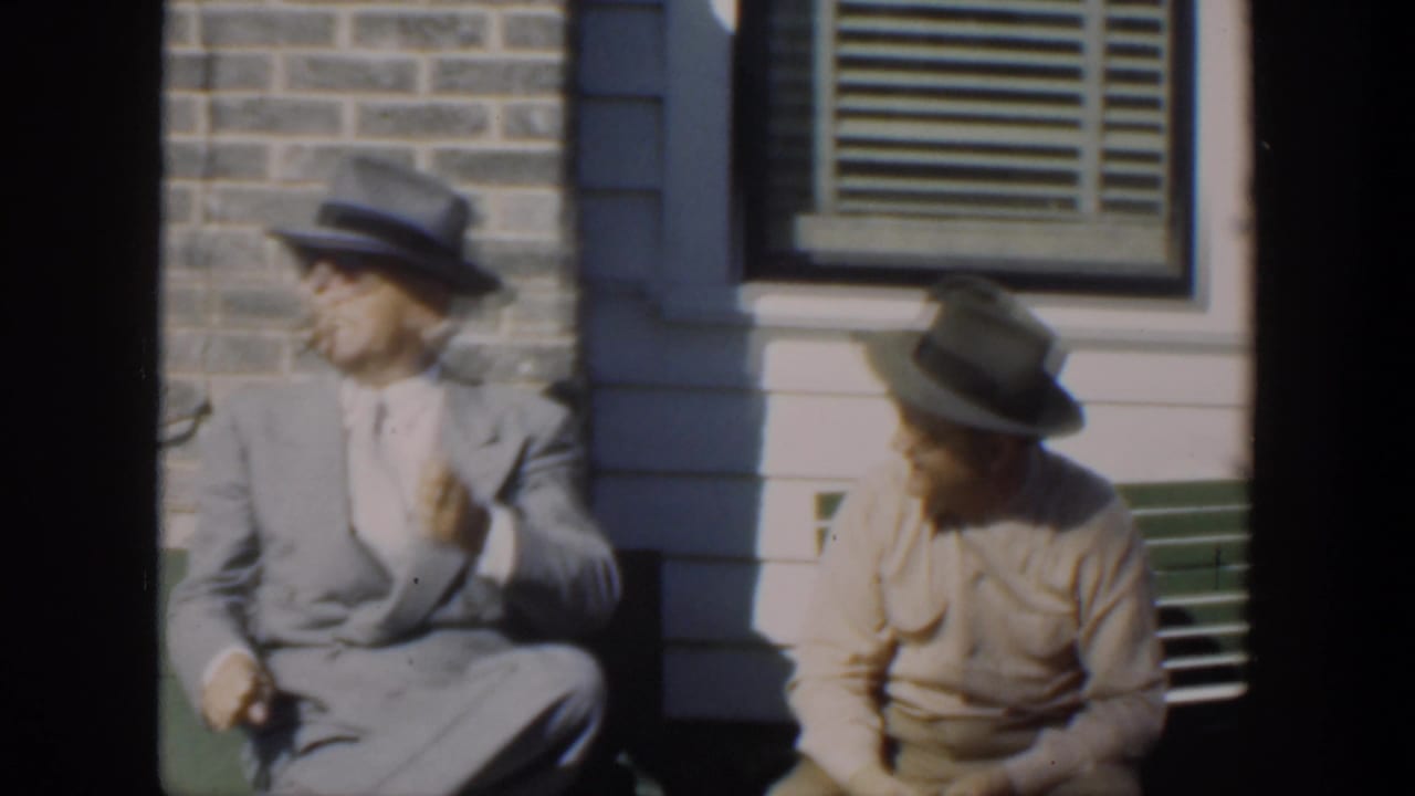 In supplied 1951 Cuba footage, two adult men in hats sit on a green bench outside a brick and clapboard house, with the man in a light suit turned to the side.