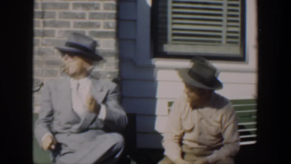 In supplied 1951 Cuba footage, two adult men in hats sit on a green bench outside a brick and clapboard house, with the man in a light suit turned to the side.