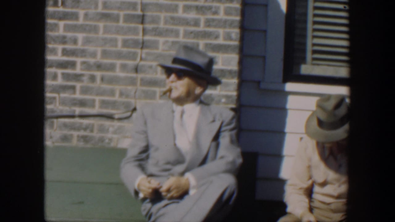 A man in a light suit and sunglasses sits on a green bench smoking a cigar.