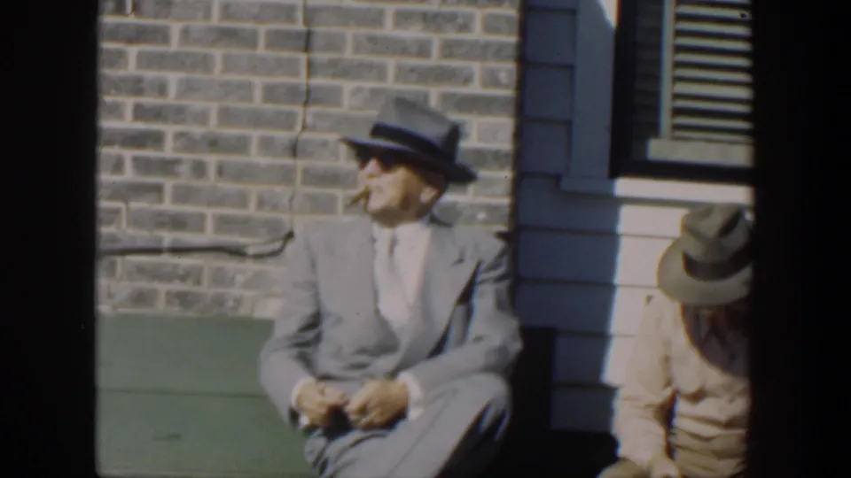 In supplied 1951 Cuba footage, a man in a light gray suit, fedora, and sunglasses sits on a green bench with a cigar while another seated man is partly visible beside him.