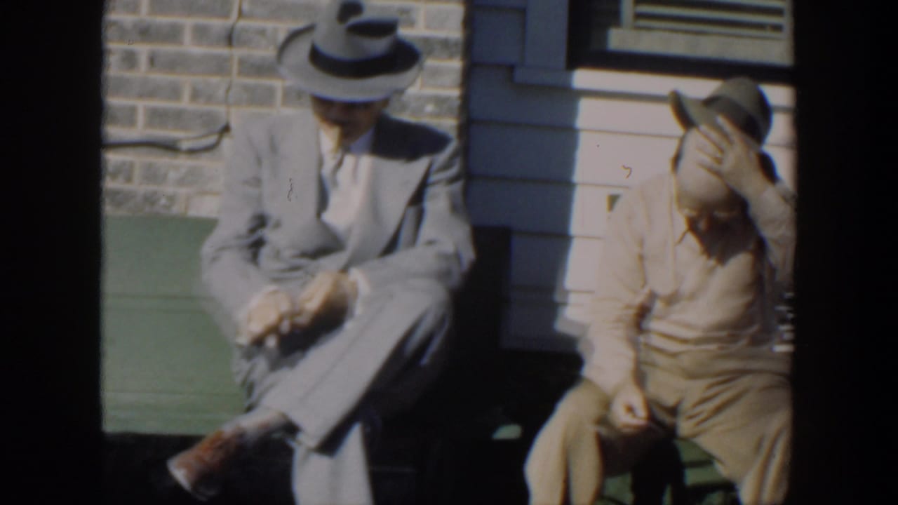 Two seated men in hats sit near a window, one lowering his head while the other looks down.