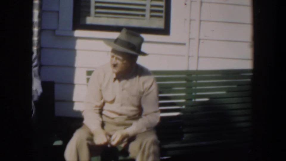 In supplied 1951 Cuba footage, an adult man in a beige shirt and fedora sits on a green wooden bench outside a clapboard wall and window, looking toward someone off frame.
