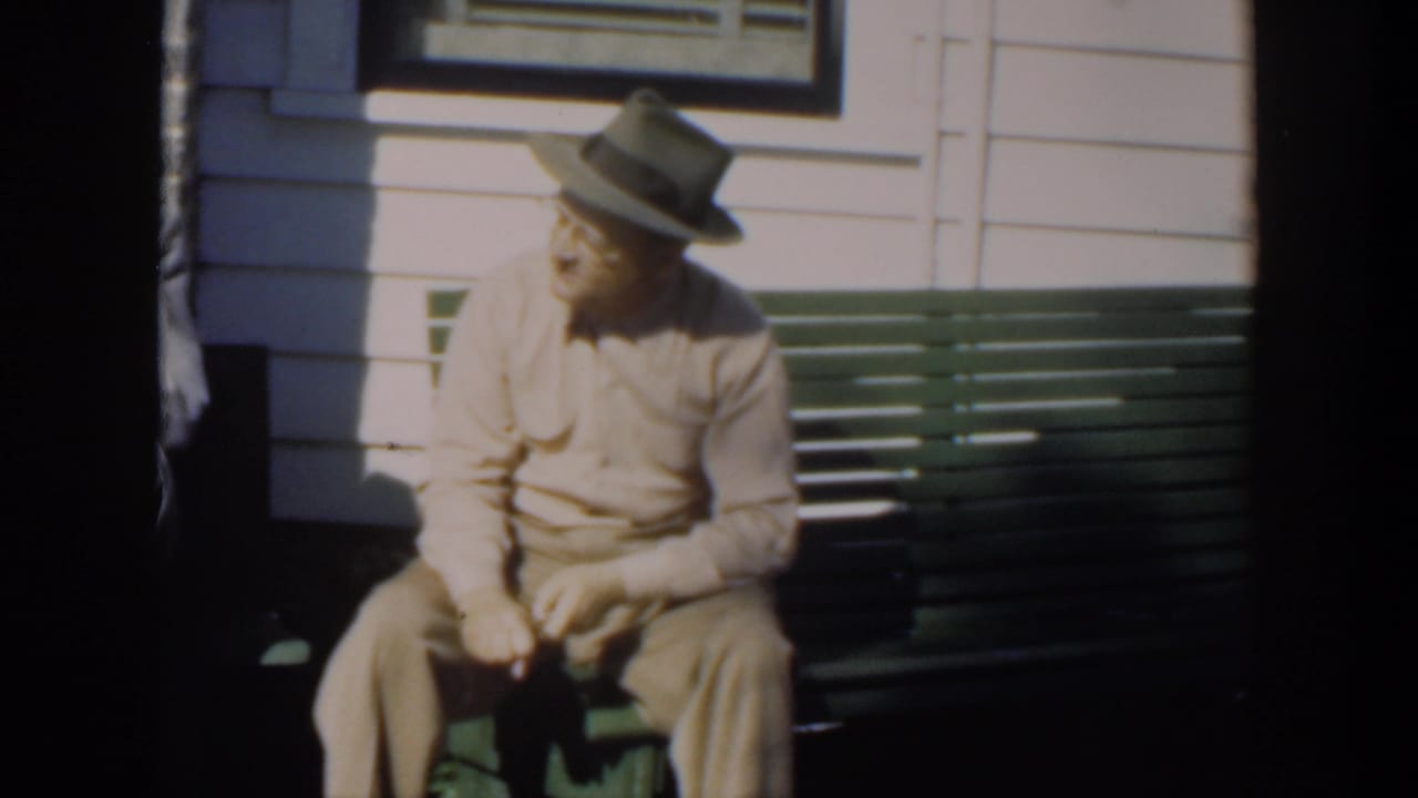Man in a fedora sitting on a green bench beside a light exterior wall.