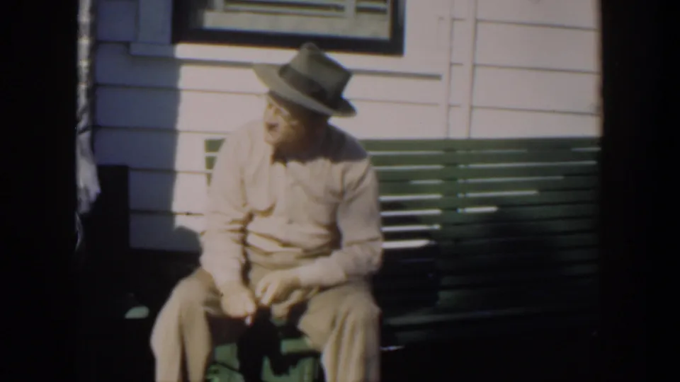 Adult man in a light shirt and brimmed hat seated on a green bench outdoors in Cuba, 1951 home-movie footage.