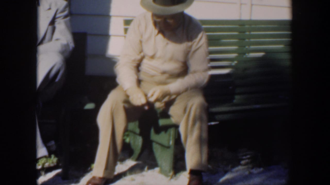 Seated man in light clothes and hat looking down while holding a small object.