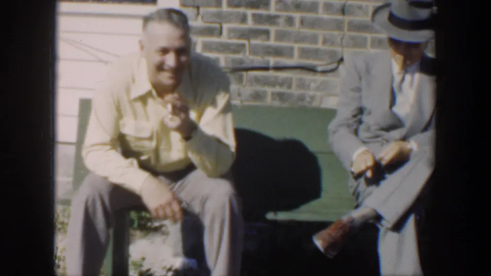 Two adult men sit on a green bench outside a brick and painted wall, with one smiling toward the camera in 1951 Cuba footage.