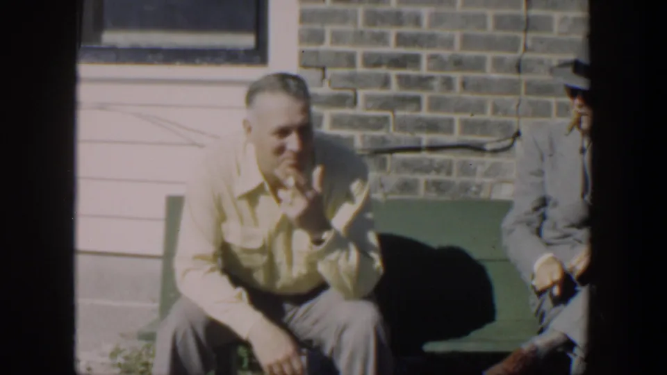 A seated man smiles on the green bench while a second suited man is partly visible beside him outdoors in Cuba, 1951.