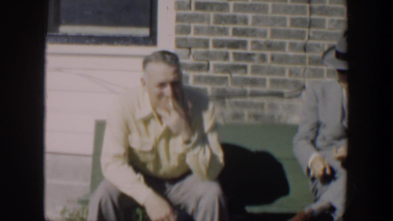 1951 Cuba home-movie frame showing adult men on a green bench outside brickwork, with one man in a light shirt smoking.
