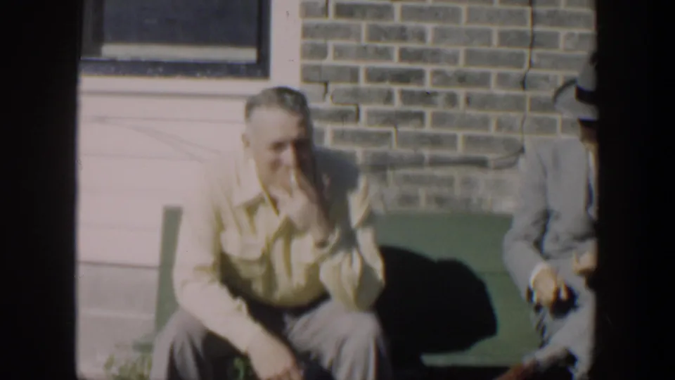 1951 Cuba home-movie frame showing adult men on a green bench outside brickwork, with one man in a light shirt smoking.