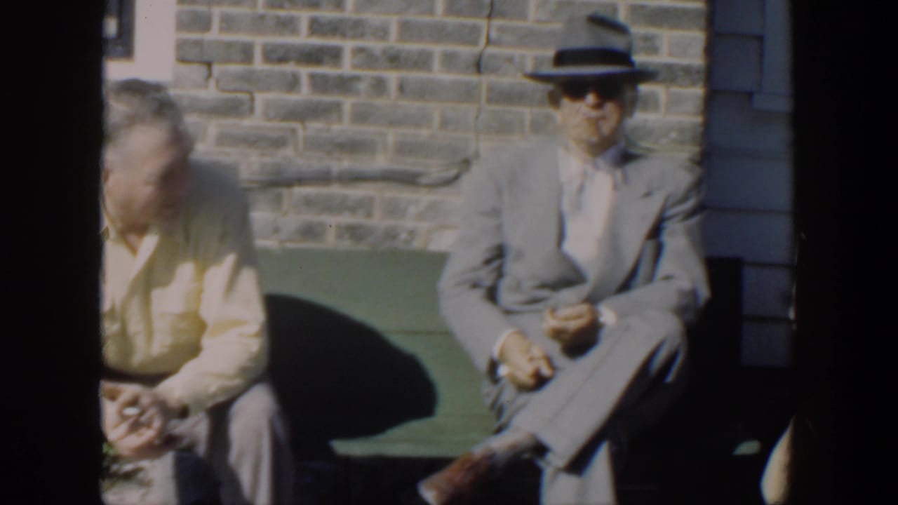 Three seated men partly divided by a dark film-frame bar, including a man in a gray suit and hat.