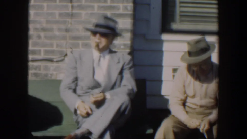1951 Cuba home-movie frame centered on a suited man in a hat seated on a green bench near brick and siding.