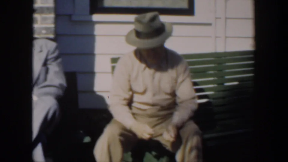 1951 Cuba frame showing a seated adult man in a light shirt and brimmed hat on a green bench in strong sunlight.