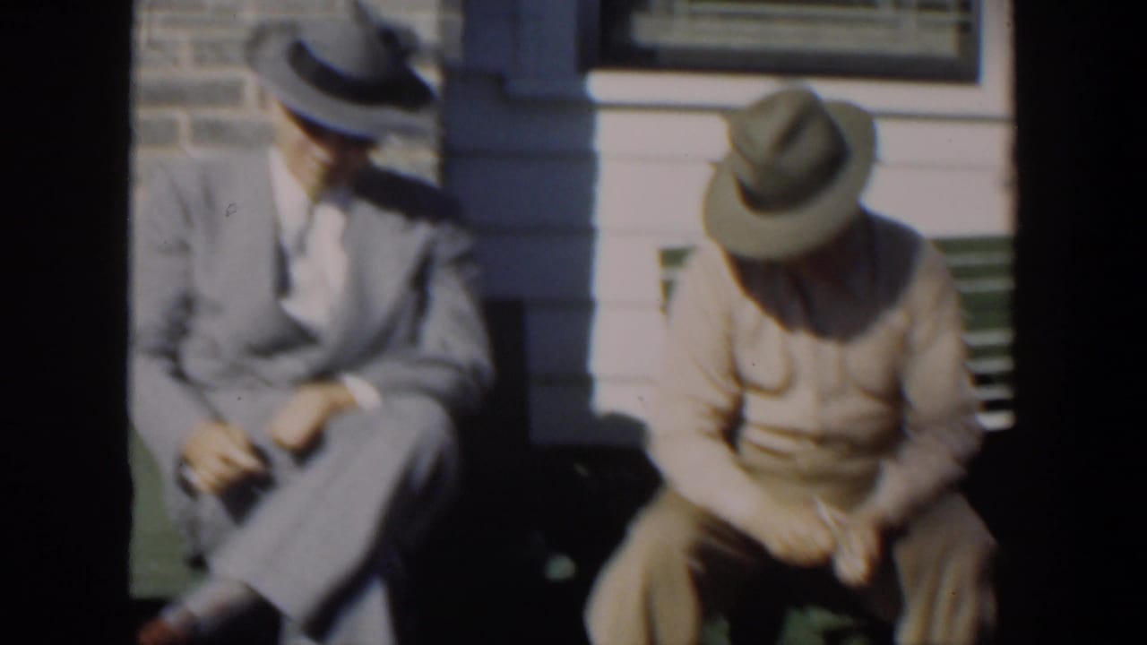 Two men in hats sit on a green bench outside a house in bright sun.