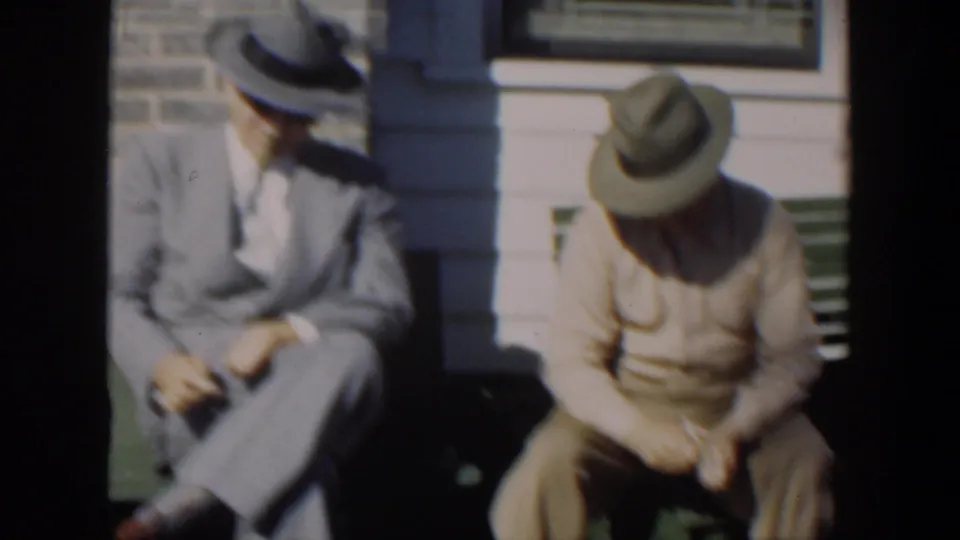 In 1951 Cuba, two older men in brimmed hats sit on a green bench outside a house, one in a suit and tie and one in beige clothing.