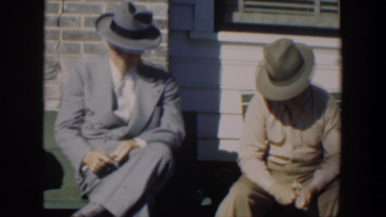 Two hatted men remain seated on a green bench, with one looking down at his hands.