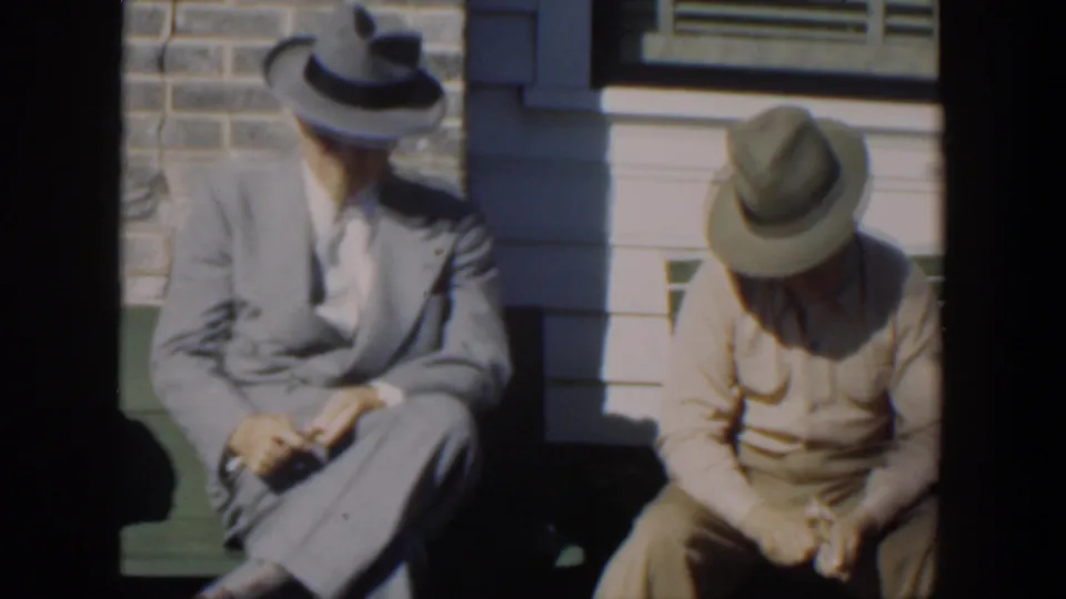 Two men sit outside on a green bench in Cuba, the suited man turned slightly toward the other as the man in beige looks down at his hands.