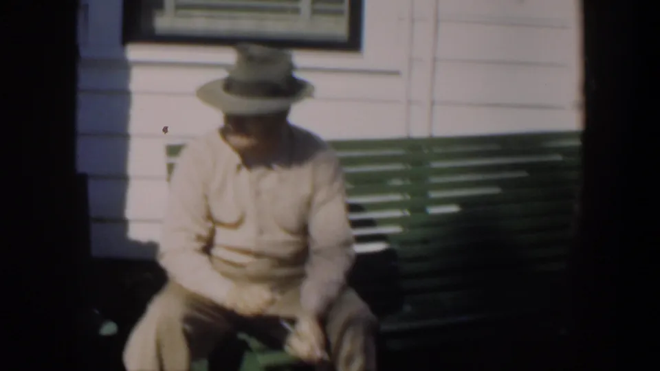 A hatted man in beige shirt and trousers sits on a green bench outside a Cuban house, looking downward with his hands near his lap.