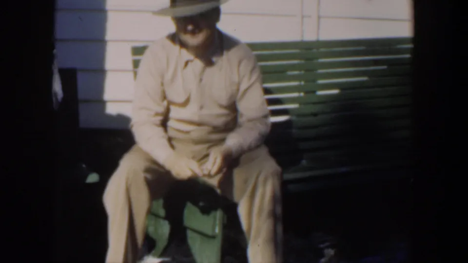A man in beige clothing and a brimmed hat sits on a green bench in Cuba, appearing to handle or carve a small object in his hands.