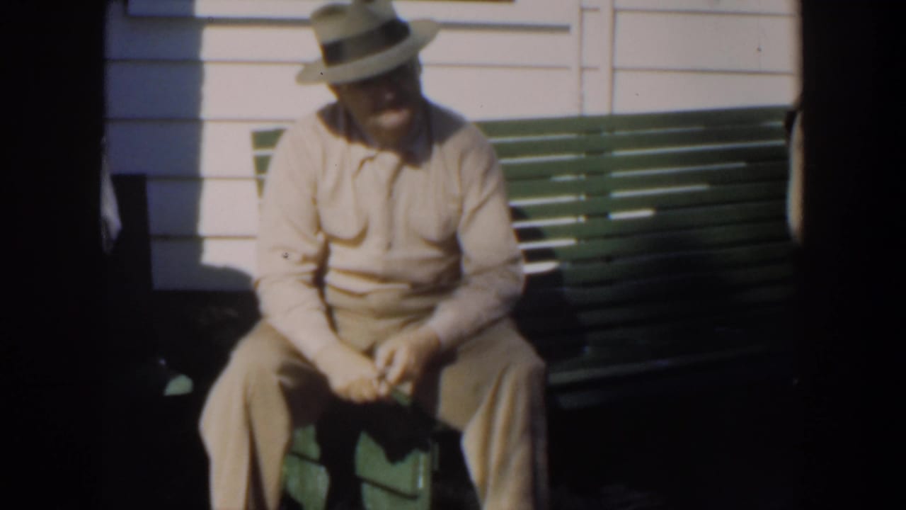 Man in a light suit and hat sits on a green bench outside a clapboard house.