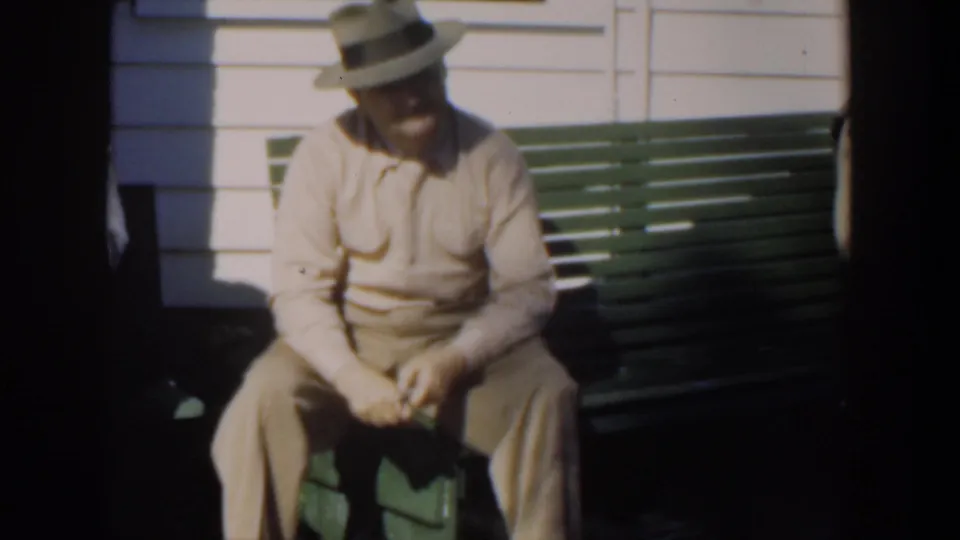 1951 Cuba home-movie frame with a man in a light suit and grey hat seated on a green bench outside a white clapboard house.