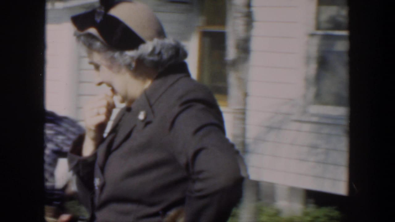 Older woman in a dark coat and hat stands near a bench outside a house.