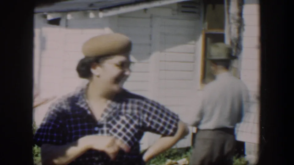 A smiling woman in a plaid shirt and beret stands near a clapboard house while a man is partly visible in the background.