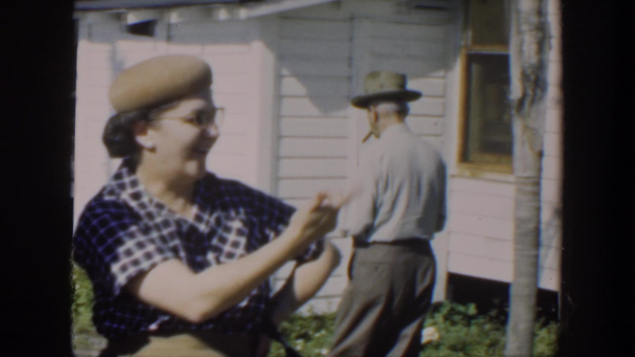 Woman in plaid dress and beige beret laughs outside a clapboard house as a man walks away behind her.