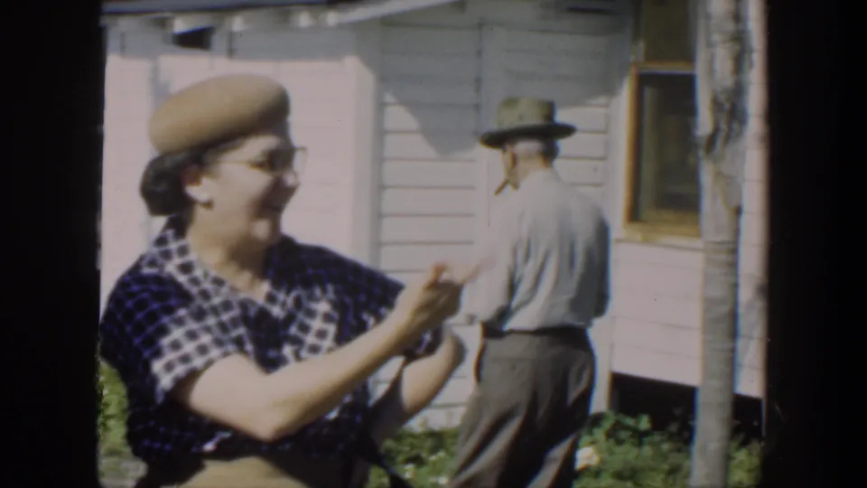 In 1951 Cuba home-movie footage, a woman in a plaid dress and beige beret laughs near a white clapboard house while a hat-wearing man turns away.