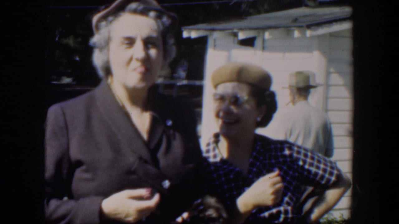 Two women stand outdoors near a clapboard house, one in a dark suit and one in a plaid dress and beret.