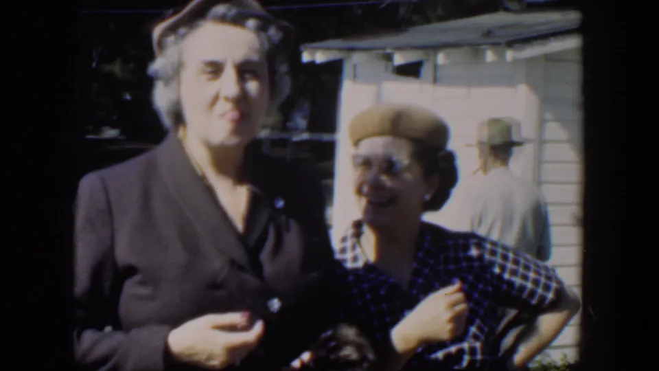 Two women in 1951 Cuba stand outside a clapboard house, one wearing a dark dress suit and the other smiling in a plaid dress and beige beret.