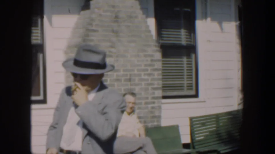 A man in a gray suit and fedora smokes beside a green bench in a 1951 Cuba home movie, with another seated man near the house wall.