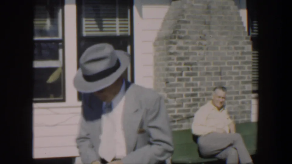 A suited man in a fedora stands near a green bench outside a house in 1951 Cuba, with another man seated against the brick wall behind him.