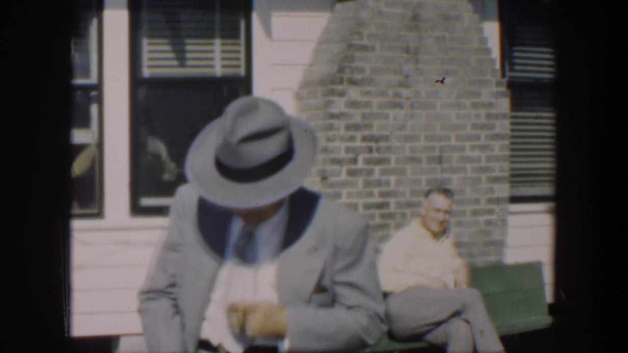 Man in a hat looks down near a white house while another adult sits on a bench behind him.