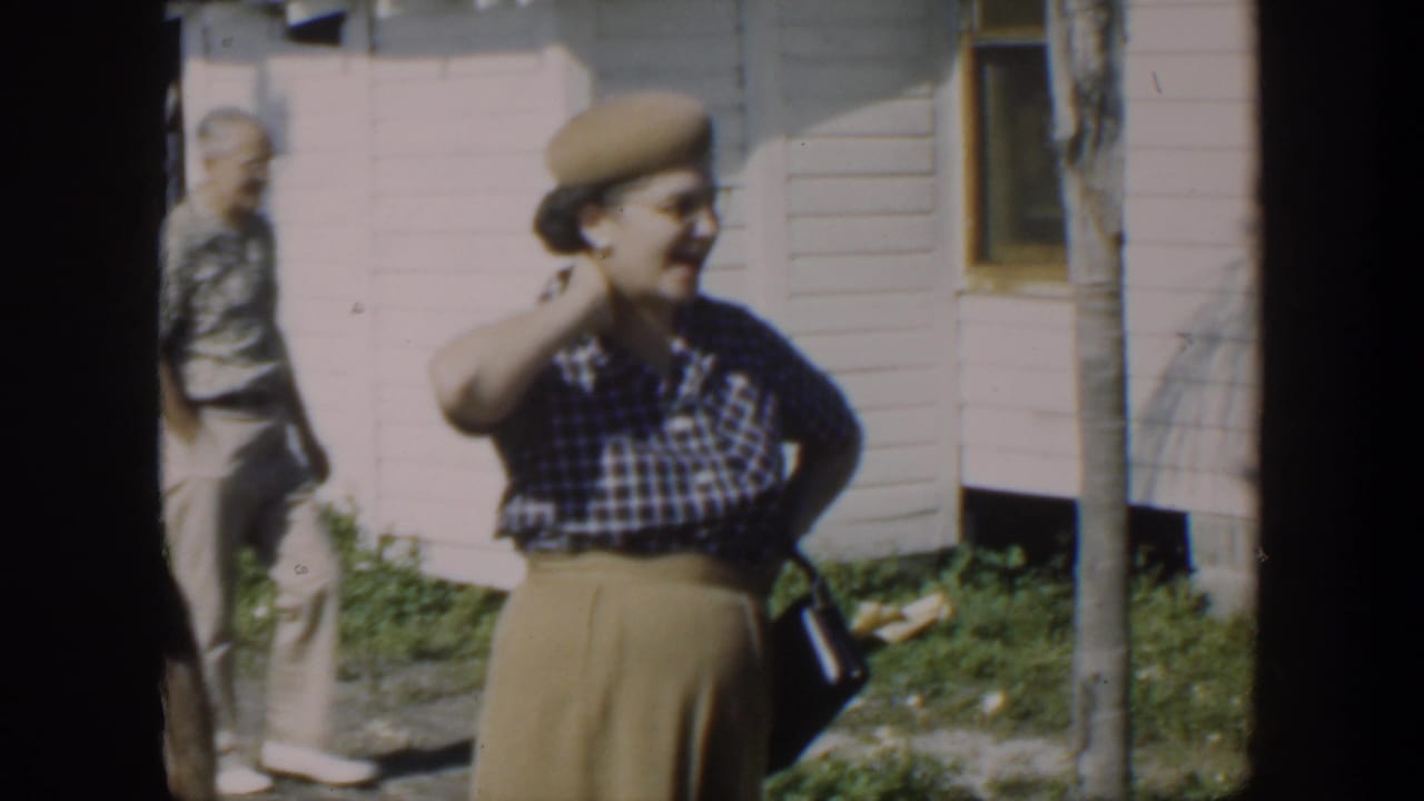 Woman in a beret and checked blouse adjusts her hair outside a white house as a man walks behind her.