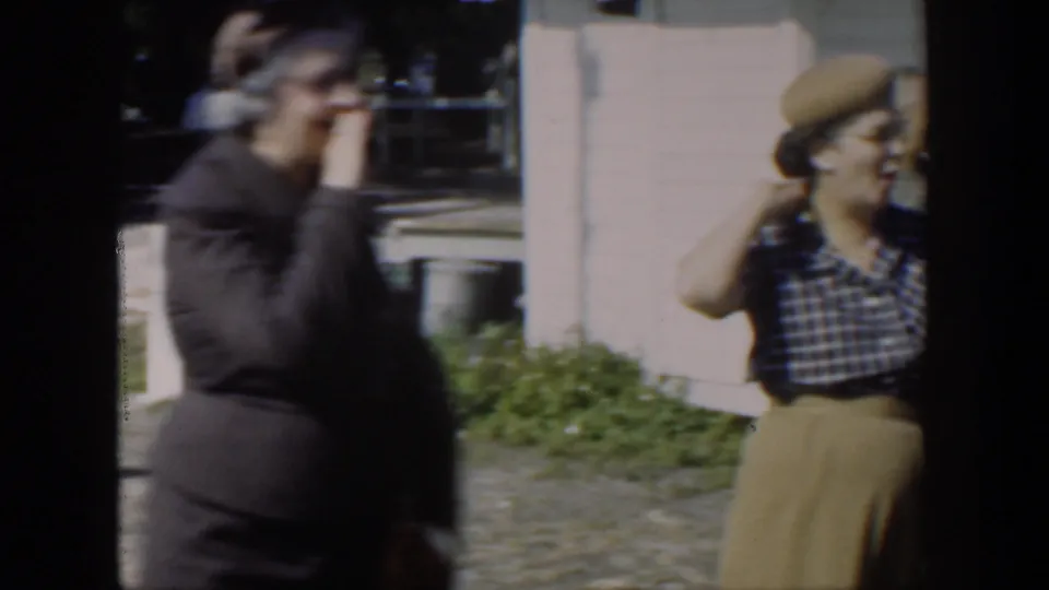 In supported 1951 Cuba footage, two adult women stand outside a clapboard house, one in dark clothing and one wearing a beret and checked blouse.