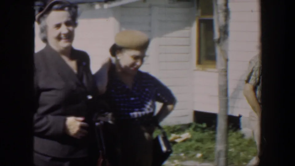 In supported 1951 Cuba footage, a smiling woman in a dark suit stands beside a woman in a beret outside a sunlit clapboard house.