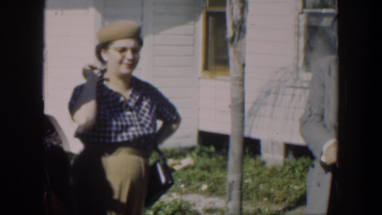 Woman in a checked dress and beige hat smiles outside a light-colored house.