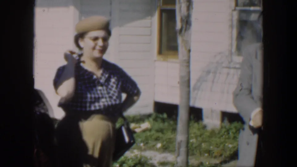 In Cuba 1951 home-movie footage, a woman in a checked dress and beige hat stands smiling near a house exterior.
