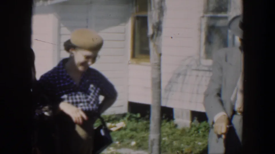 A woman in a checked dress and hat stands outside a Cuban house as the home-movie camera catches partial figures and dark film edges.